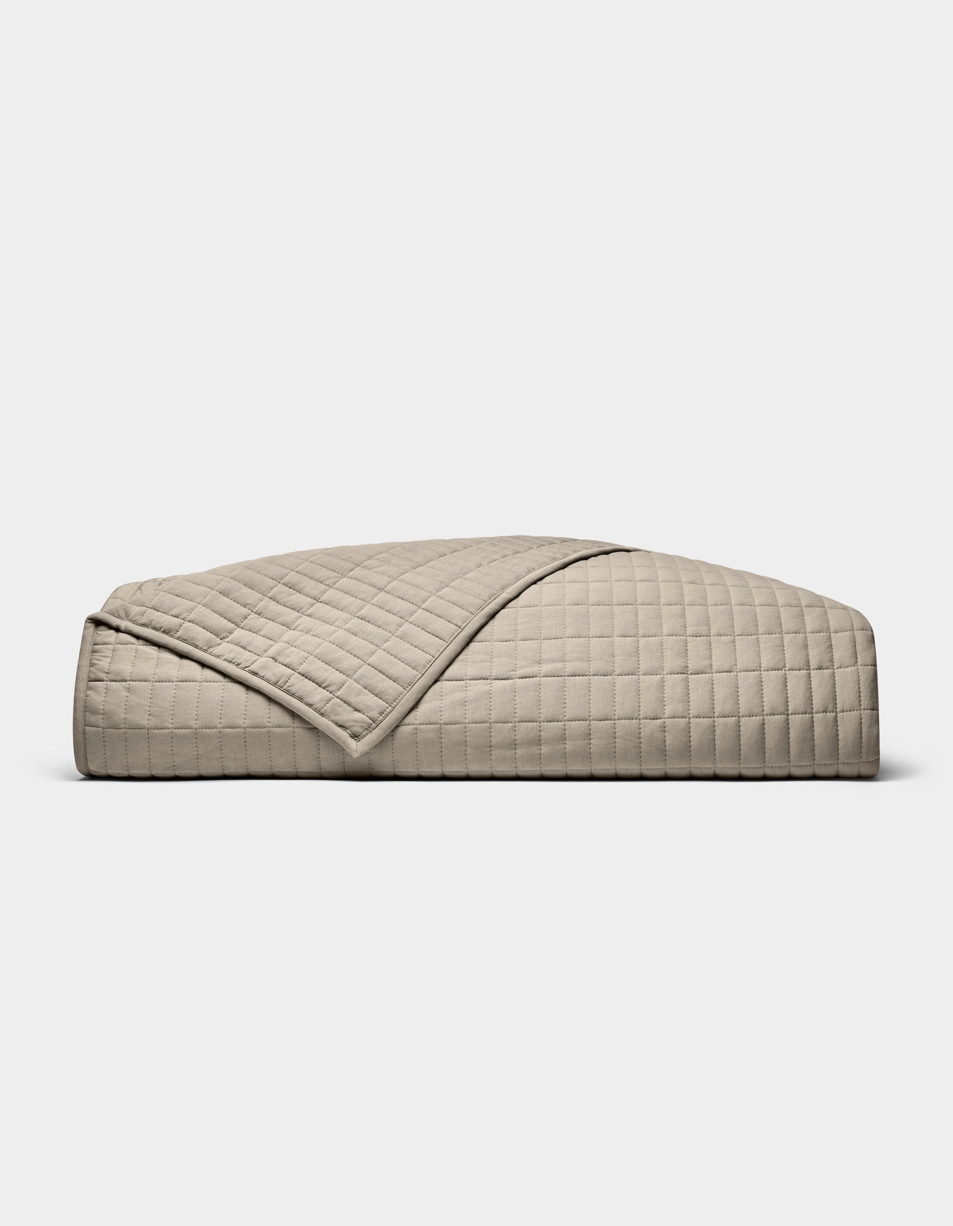 The Cozy Earth Bamboo Jersey Quilt, featuring a beige, quilted grid pattern, is neatly folded on a white background. A turned-down corner highlights its soft, luxurious texture.|Color:Driftwood