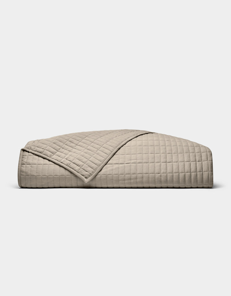 The Cozy Earth Bamboo Jersey Quilt, featuring a beige, quilted grid pattern, is neatly folded on a white background. A turned-down corner highlights its soft, luxurious texture.