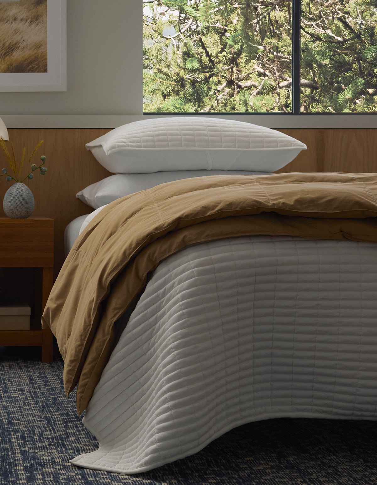 A bed styled with Cozy Earth's Down Alternative Throw features white quilted bedding, a tan comforter, and four stacked white pillows. Next to it sits a wooden nightstand with a vase and lamp, while green tree branches are visible through the window.