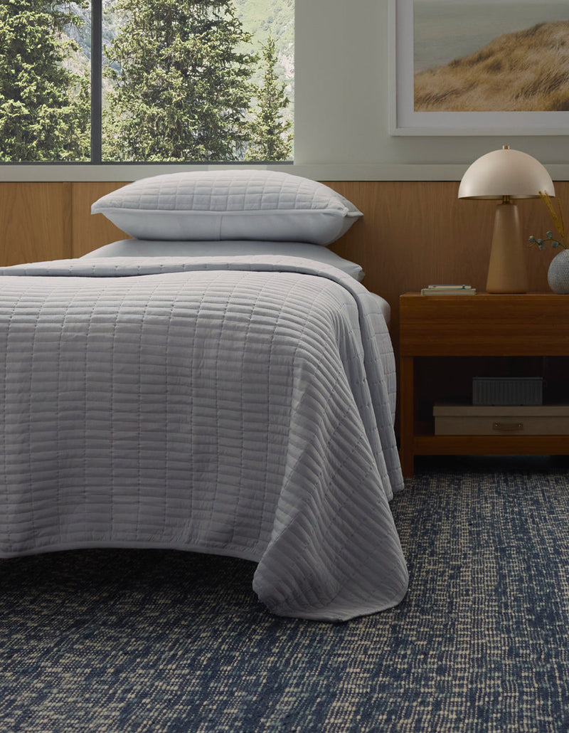 A bed with the Cozy Earth Bamboo Jersey Quilt in light gray and matching pillow is next to a wooden nightstand with a lamp, all atop a blue patterned rug. A large window and a framed landscape art piece are visible in the background.