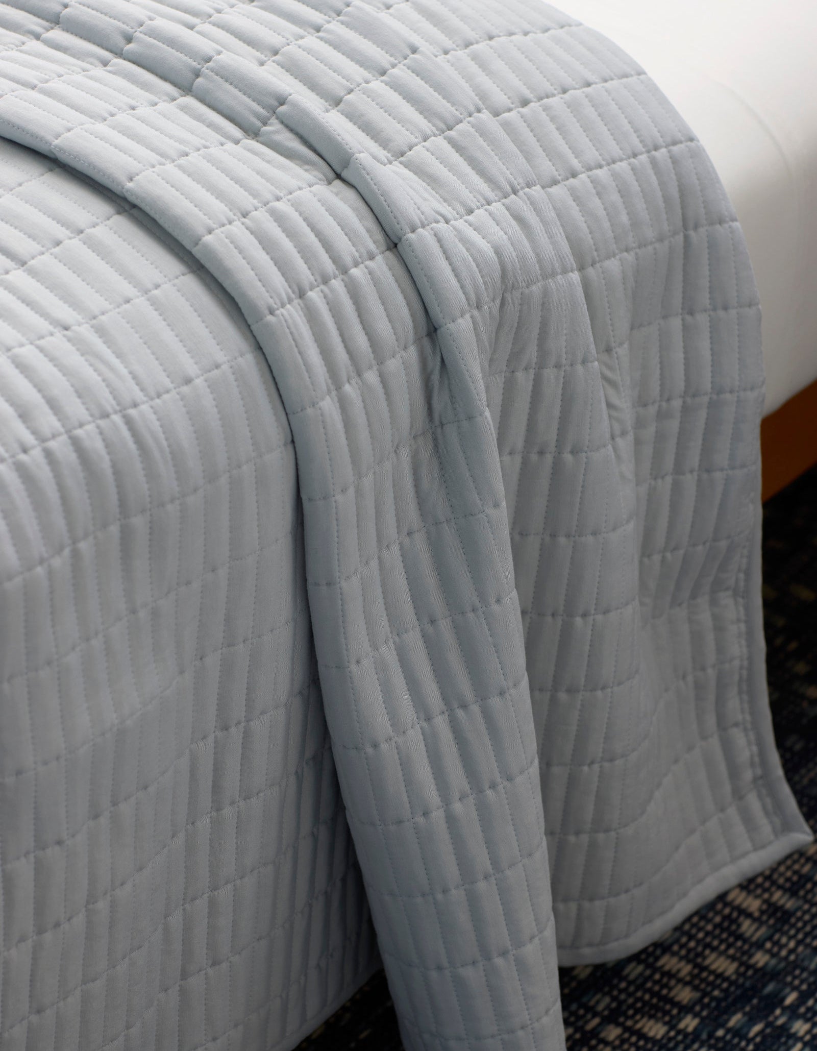 The Cozy Earth Bamboo Jersey Quilt, featuring a light gray quilted grid pattern, is draped neatly over a bed, partially hanging toward a blue patterned carpet below.|Color:Shore