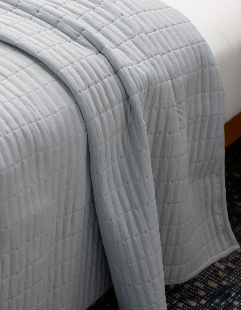 The Cozy Earth Bamboo Jersey Quilt, featuring a light gray quilted grid pattern, is draped neatly over a bed, partially hanging toward a blue patterned carpet below.
