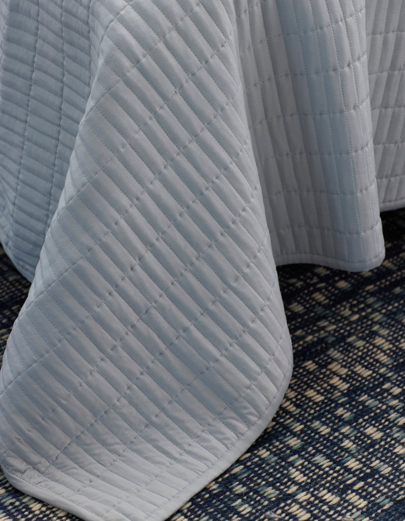 A close-up of the Cozy Earth Bamboo Jersey Quilt in light gray, draped over a surface, with a dark blue and cream patterned carpet visible beneath.