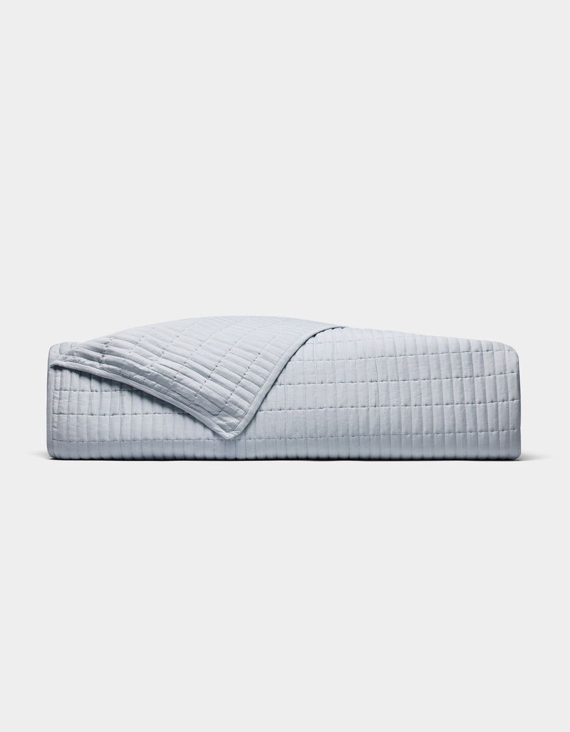 A neatly made bed displays the Cozy Earth Bamboo Jersey Quilt in light grey with a matching pillow, one quilt corner folded back, all set against a plain white background.