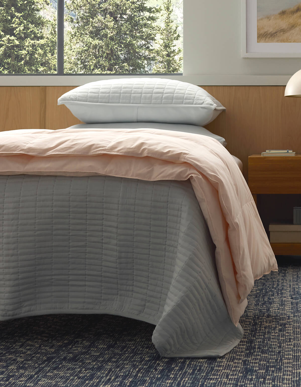 A bed features the Cozy Earth Down Alternative Throw in light gray, a pale pink comforter, and two stacked pillows. Nearby is a wooden nightstand, a blue patterned carpet, and a window with views of green trees.
