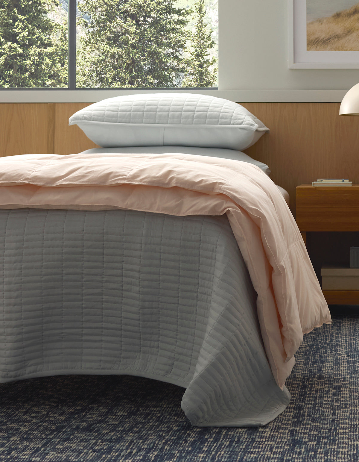 A modern bedroom with wood paneling features a neatly made bed topped with Cozy Earth's Down Alternative Throw, a gray quilted bedspread, light pink comforter, and two stacked gray pillows on a blue-patterned carpet.