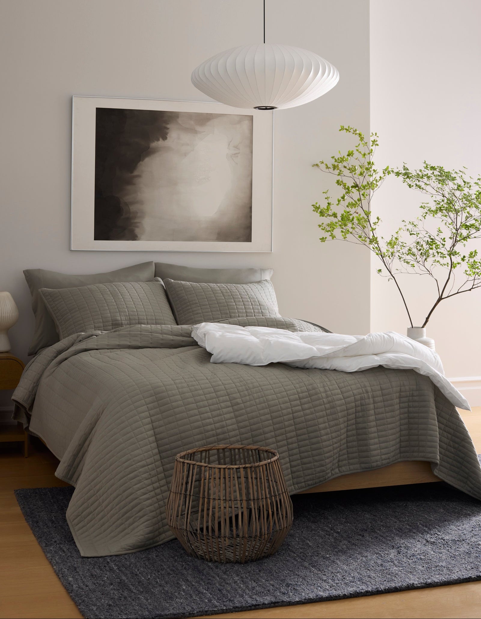 A modern bedroom features a neatly made bed with the Cozy Earth Bamboo Jersey Quilt, a large abstract artwork above the headboard, a paper lantern ceiling light, a green leafy plant, a woven basket, and a dark rug on light wood flooring.|Color:Stone