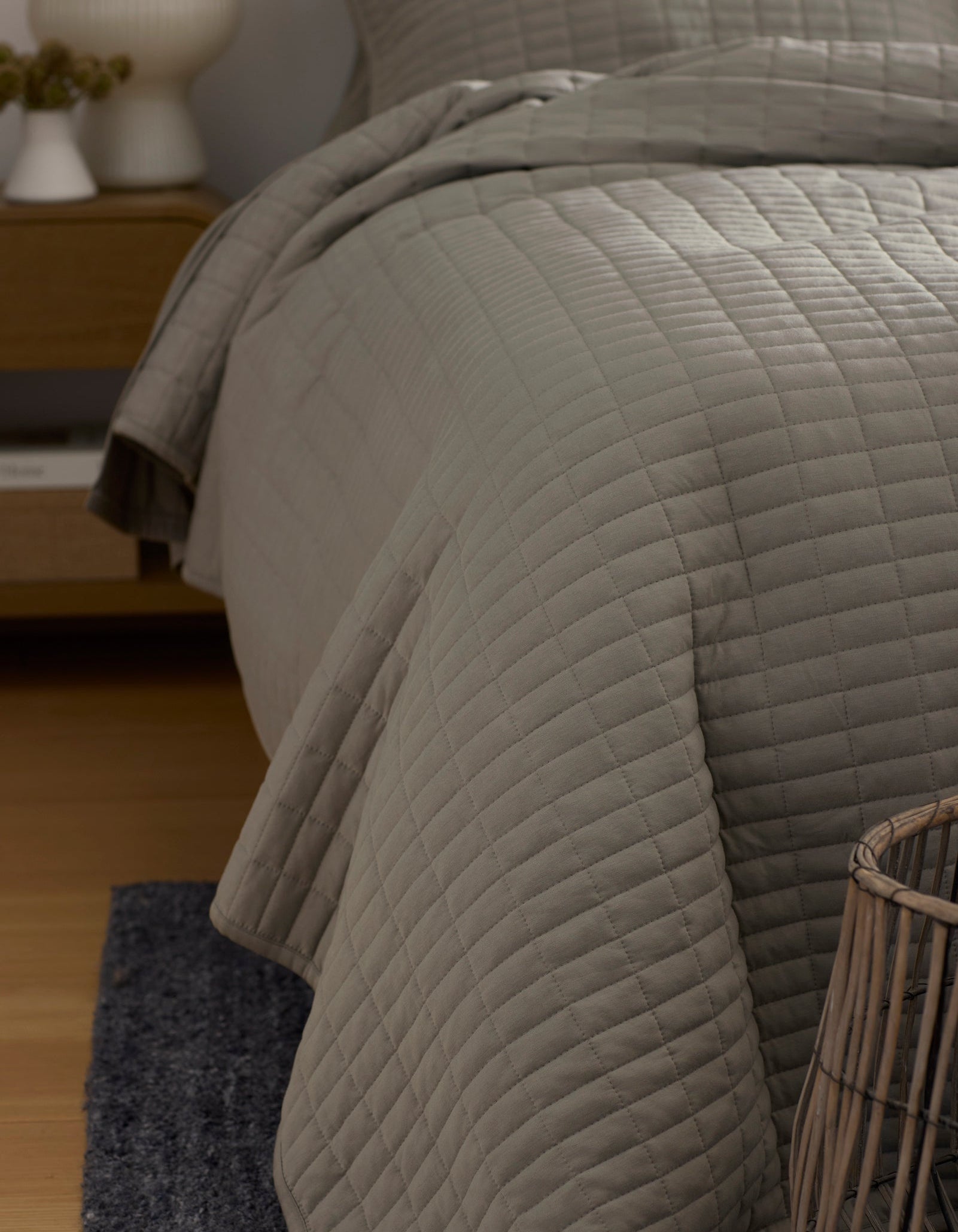 A close-up of a neatly made bed features the Cozy Earth Bamboo Jersey Quilt in light gray, draped over the side. Also visible are part of a wicker basket, a dark rug, and a wooden nightstand in this cozy bedroom corner.|Color:Stone