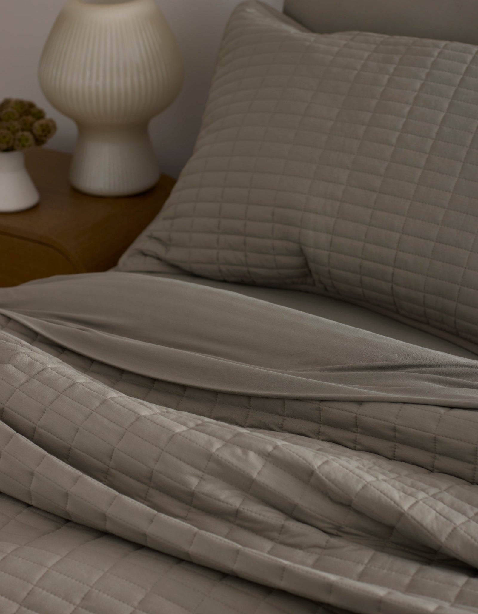 A neatly made bed features the Bamboo Jersey Quilt by Cozy Earth in taupe with matching pillows, next to a wooden nightstand topped with a white ribbed lamp and a small flower vase.