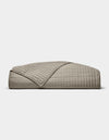 The Cozy Earth Bamboo Jersey Quilt is shown folded against a white background, with one corner turned down to display its beige color, grid pattern, and detailed stitching.|Color:Stone