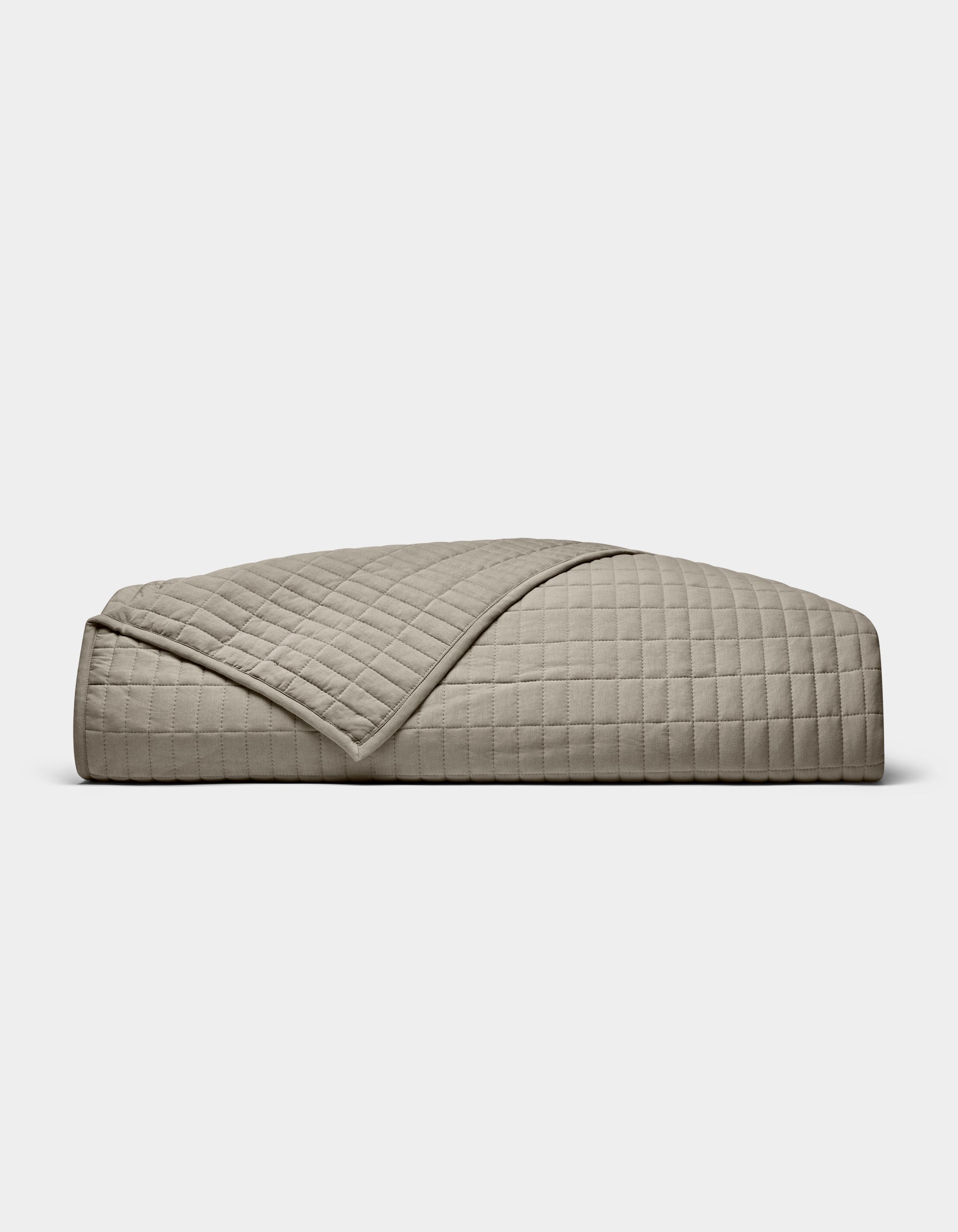 The Cozy Earth Bamboo Jersey Quilt is shown folded against a white background, with one corner turned down to display its beige color, grid pattern, and detailed stitching.|Color:Stone