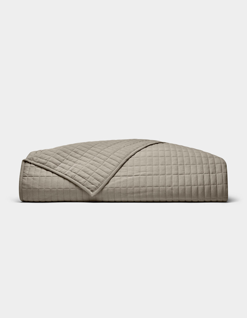 The Cozy Earth Bamboo Jersey Quilt is shown folded against a white background, with one corner turned down to display its beige color, grid pattern, and detailed stitching.