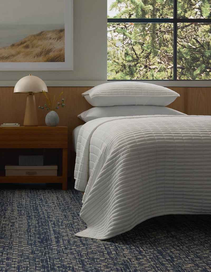 A neatly made bed features the Cozy Earth Bamboo Jersey Quilt and pillows, next to a wooden nightstand with a lamp and vase; a large window reveals green trees, while a textured blue rug covers the floor.