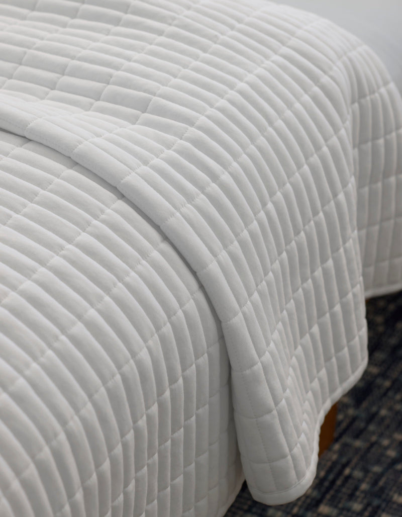 A close-up of a neatly made bed with the Cozy Earth Bamboo Jersey Quilt, featuring a white, square-patterned design, draped over the edge atop a dark patterned rug.