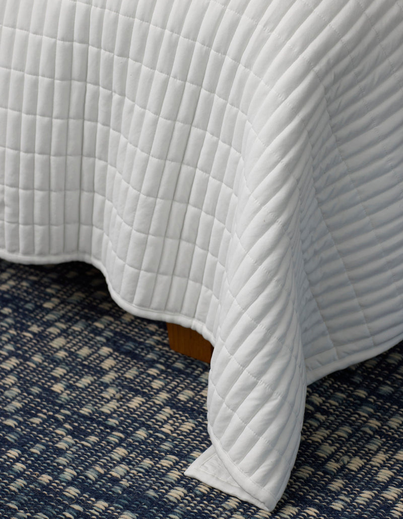 A close-up of the Cozy Earth Bamboo Jersey Quilt, featuring a white quilted grid pattern, draped over a bed with a blue and white patterned carpet underneath.