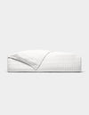 The Cozy Earth Bamboo Jersey Quilt is shown neatly made on a bed, its white, textured fabric partially folded back, photographed from the side against a plain light grey background.|Color:White