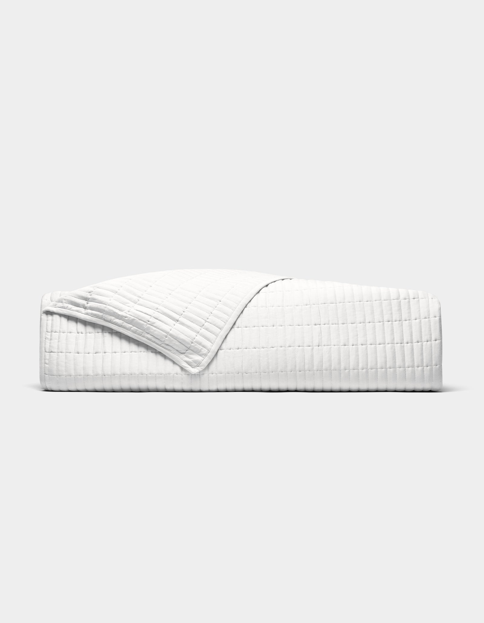 The Cozy Earth Bamboo Jersey Quilt is shown neatly made on a bed, its white, textured fabric partially folded back, photographed from the side against a plain light grey background.