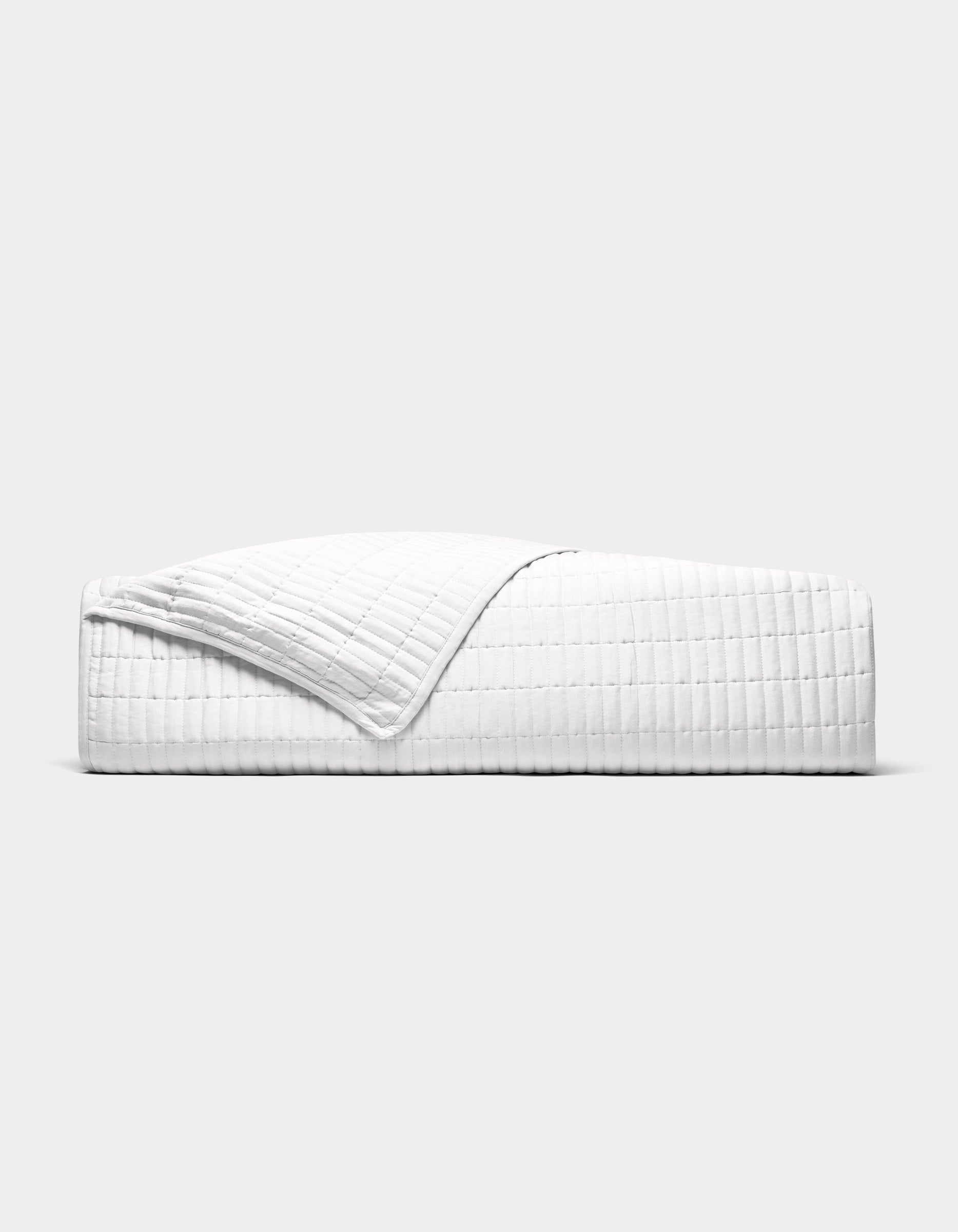 The Cozy Earth Bamboo Jersey Quilt is shown neatly made on a bed, its white, textured fabric partially folded back, photographed from the side against a plain light grey background.|Color:White