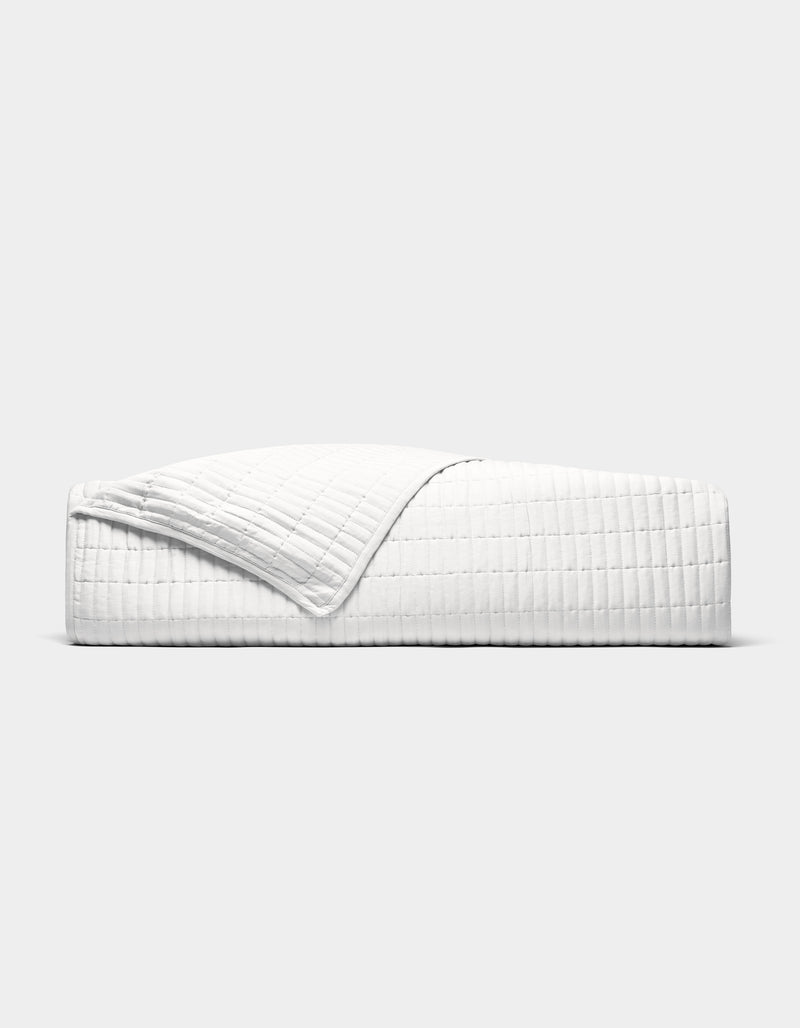 The Cozy Earth Bamboo Jersey Quilt is shown neatly made on a bed, its white, textured fabric partially folded back, photographed from the side against a plain light grey background.