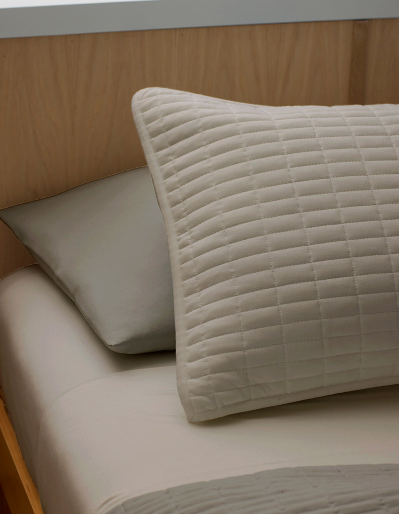 A close-up of a neatly made bed features two pillows resting against a light wooden headboard: a Bamboo Jersey Quilted Sham by Cozy Earth in beige in front, and a plain gray pillow behind.