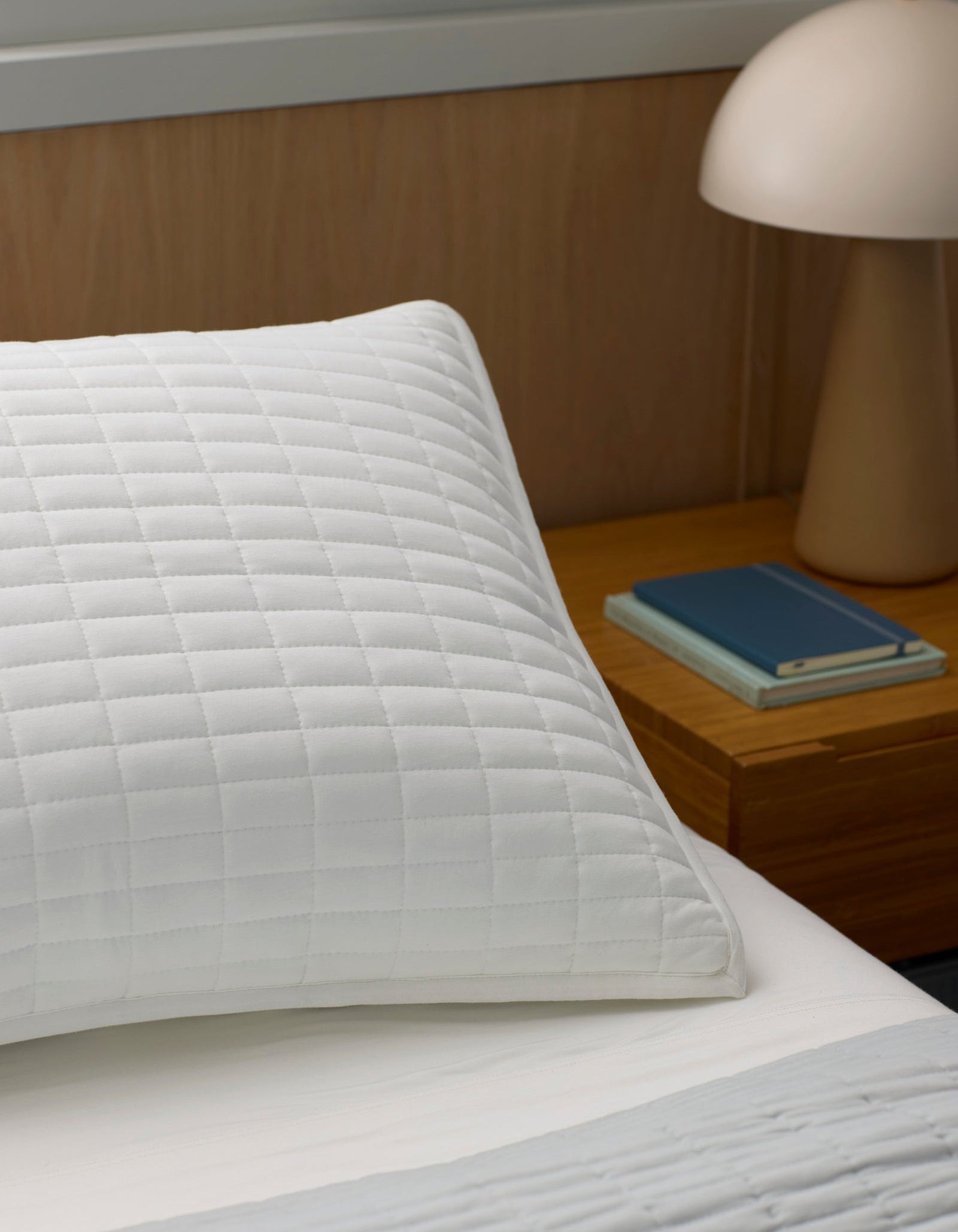 A close-up of a white Bamboo Jersey Quilted Sham by Cozy Earth, resting on a bed beside a wooden nightstand with a beige lamp and two neatly stacked closed books.