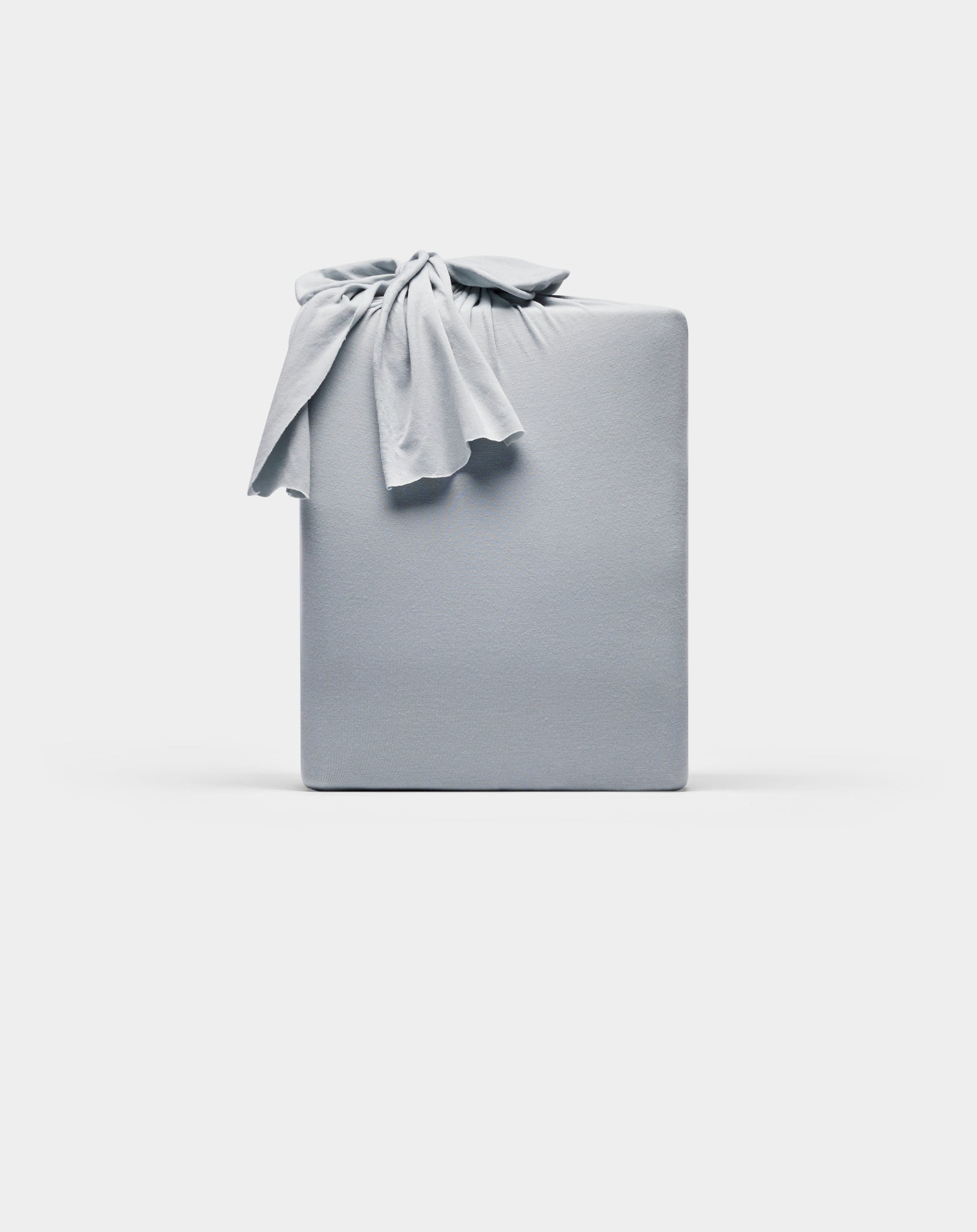 A rectangular Bamboo Jersey Sheet Set by Cozy Earth, wrapped in smooth, light gray fabric with a knot on top, displayed against a plain white background.
