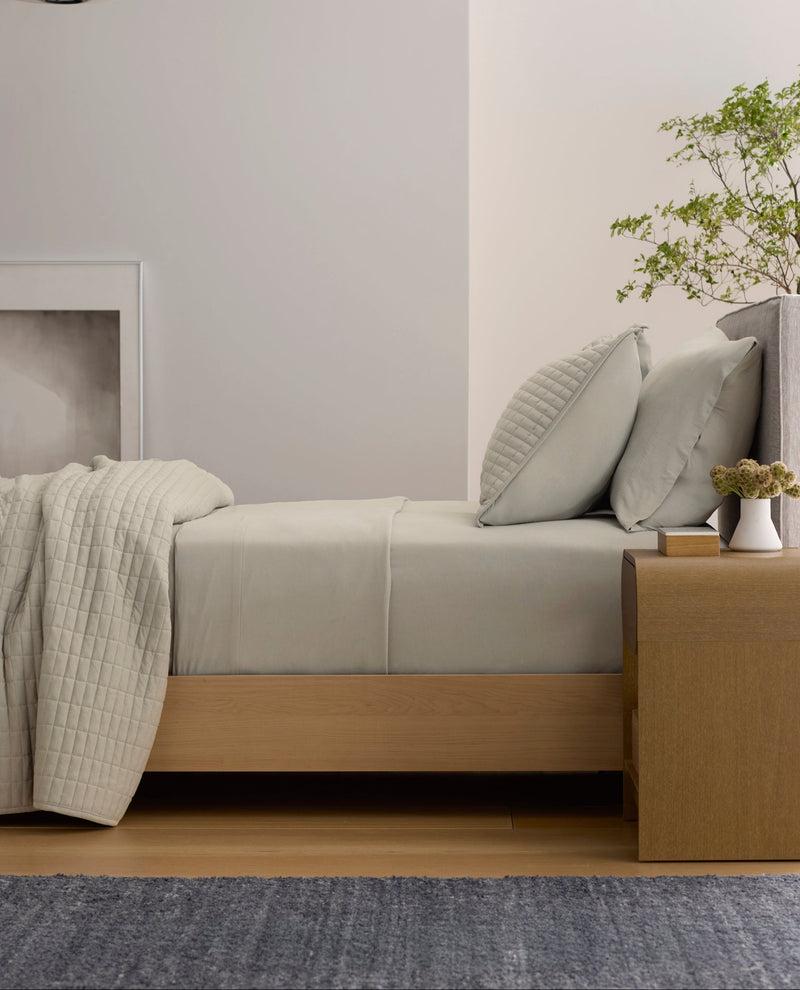 A minimalist room features a neatly made bed with Cozy Earth's Bamboo Jersey Flat Sheet in light gray, paired with pillows. Next to it is a wooden nightstand holding a small plant and vase, while a framed picture and pendant lamp complete the scene.