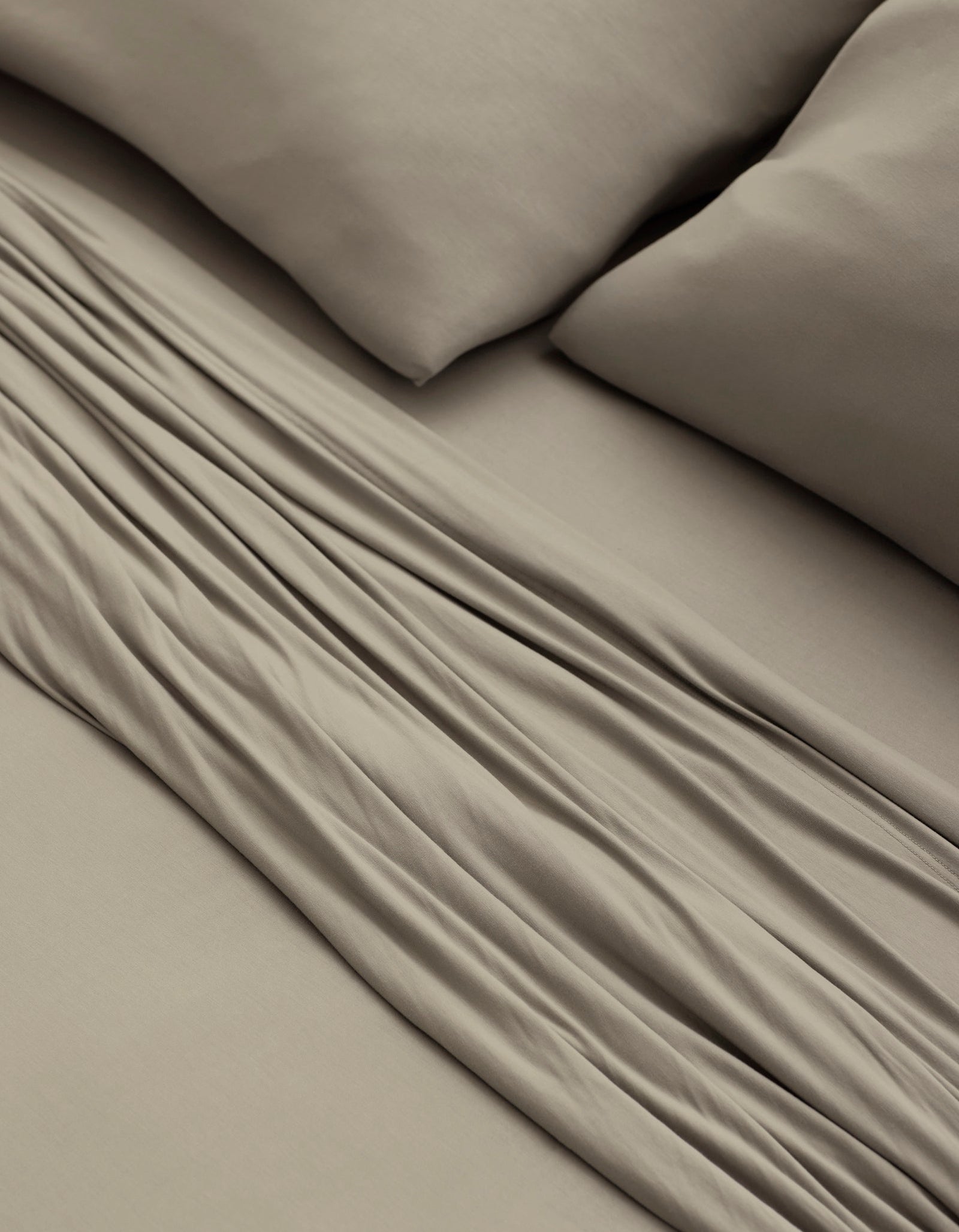 A close-up of a bed made with Cozy Earth’s Bamboo Jersey Sheet Set in taupe, featuring two matching pillows and showcasing the soft fabric folds and smooth texture.