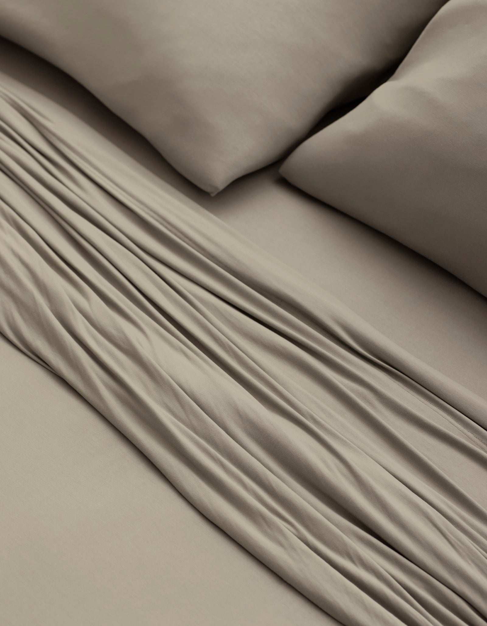 A close-up of a bed made with Cozy Earth’s Bamboo Jersey Sheet Set in taupe, featuring two matching pillows and showcasing the soft fabric folds and smooth texture.