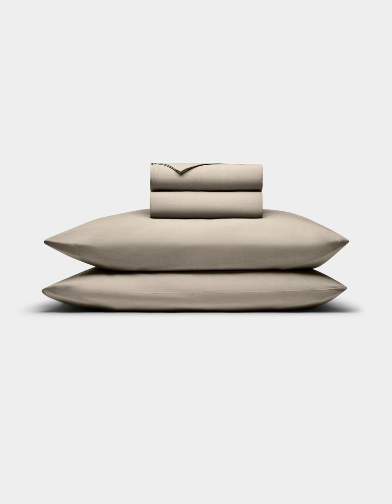 The Bamboo Jersey Sheet Set by Cozy Earth is displayed on a plain white background, featuring a neatly folded beige set with two stacked pillowcases, a fitted sheet, and a flat sheet.