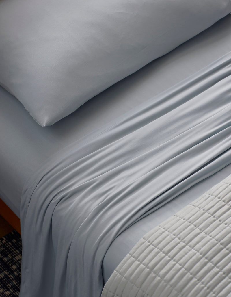 A close-up of a neatly made bed features the Cozy Earth Bamboo Jersey Sheet Set in light gray, with silky sheets and pillowcase. A textured, light quilt is folded at the foot, highlighting the smooth, soft bedding.