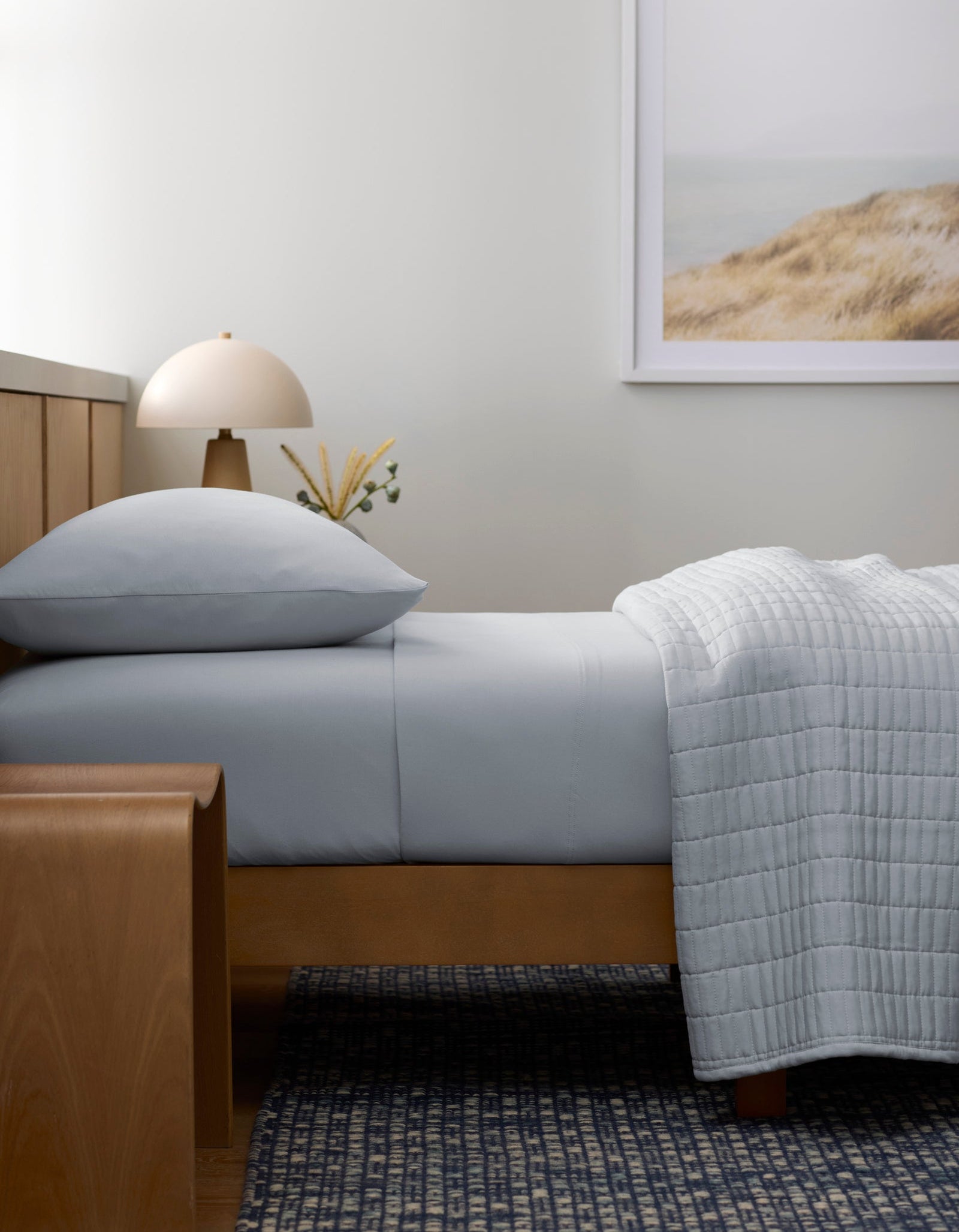 A minimalist bedroom features a neatly made bed with Cozy Earth's Bamboo Jersey Flat Sheet in light gray, a wooden bench, a small lamp, and a landscape dunes photo; natural light enhances the serene, neutral space.