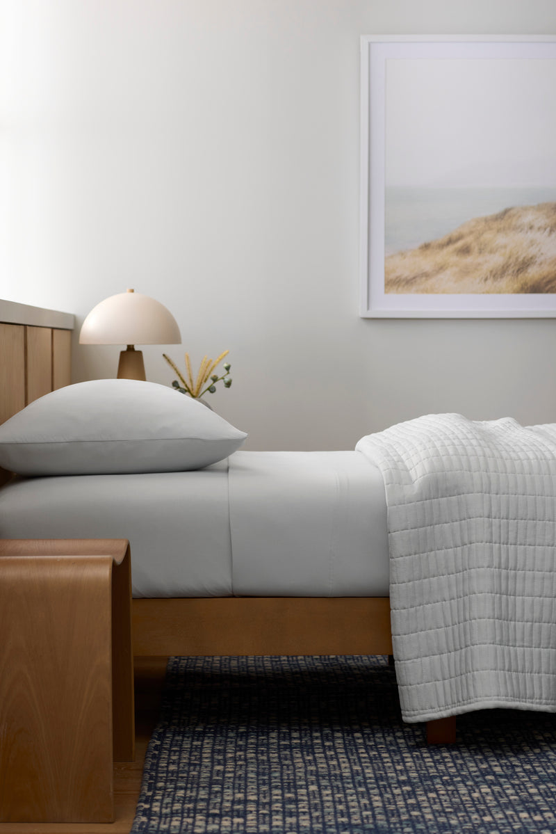 A modern, minimal bedroom features the Cozy Earth Bamboo Jersey Fitted Sheet in light gray, a wooden bench, a small round table lamp, a framed beach landscape on the wall, and a blue patterned rug on the floor.