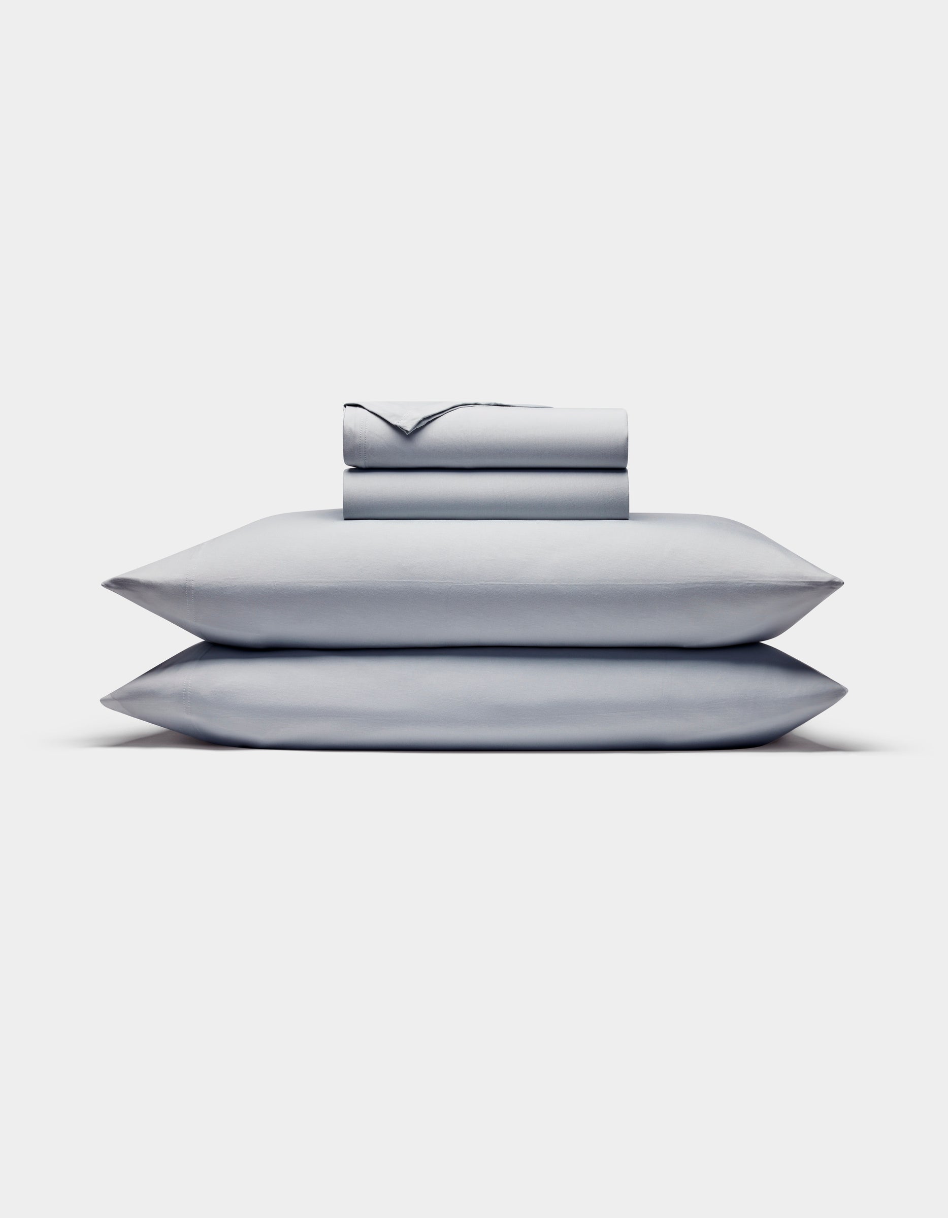 The Cozy Earth Bamboo Jersey Sheet Set is shown neatly stacked with two pillows at the bottom and folded light gray sheets and pillowcases on top, all set against a plain white background.|Color:Shore