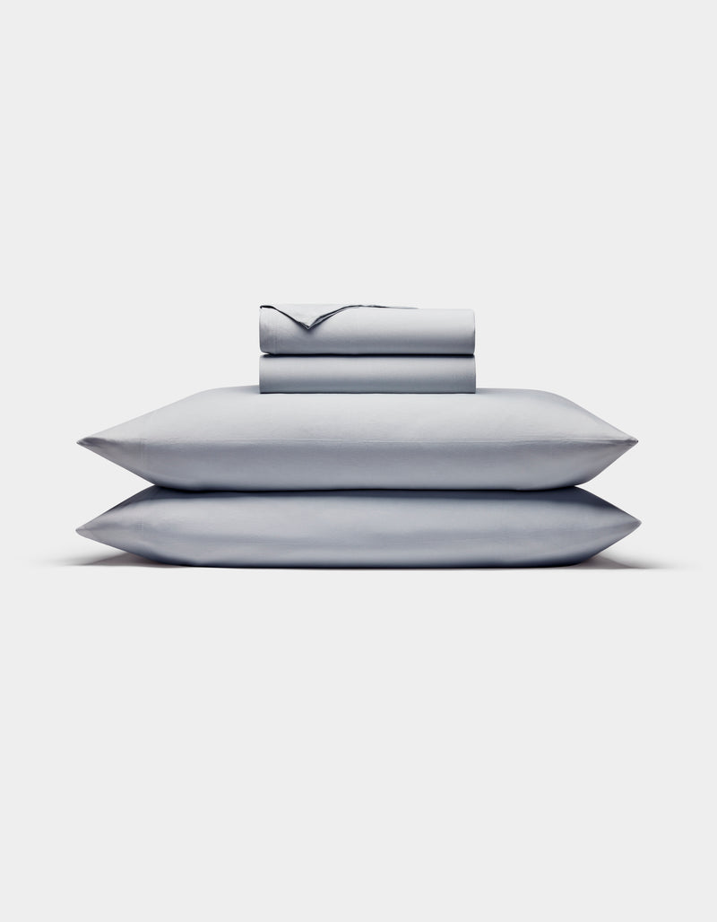 The Cozy Earth Bamboo Jersey Sheet Set is shown neatly stacked with two pillows at the bottom and folded light gray sheets and pillowcases on top, all set against a plain white background.