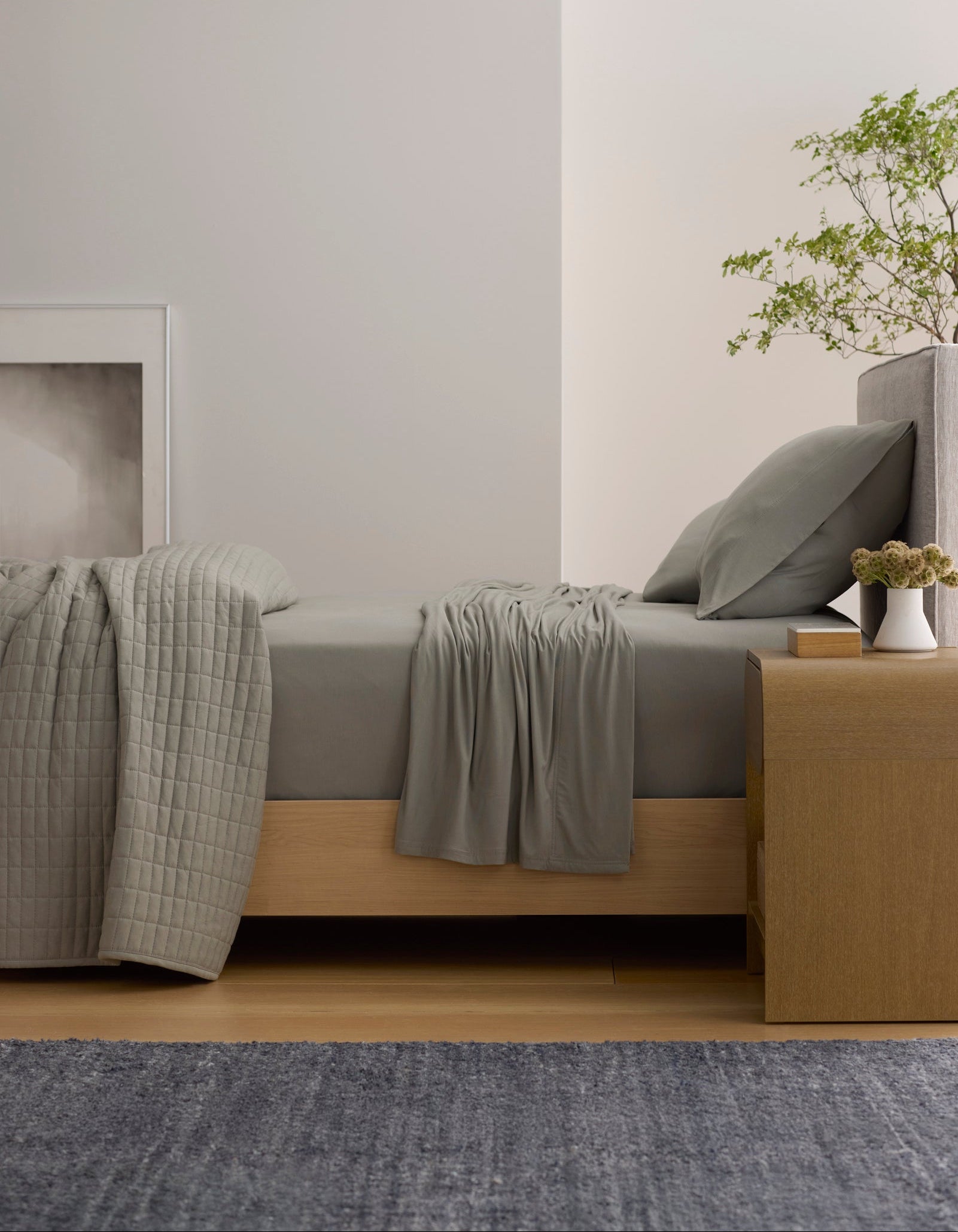 A minimalist bedroom features a Cozy Earth Bamboo Jersey Sheet Set in gray on a wooden bed with matching pillows. A wooden nightstand holds a small plant and flowers, while large abstract art and a modern pendant light add style.
