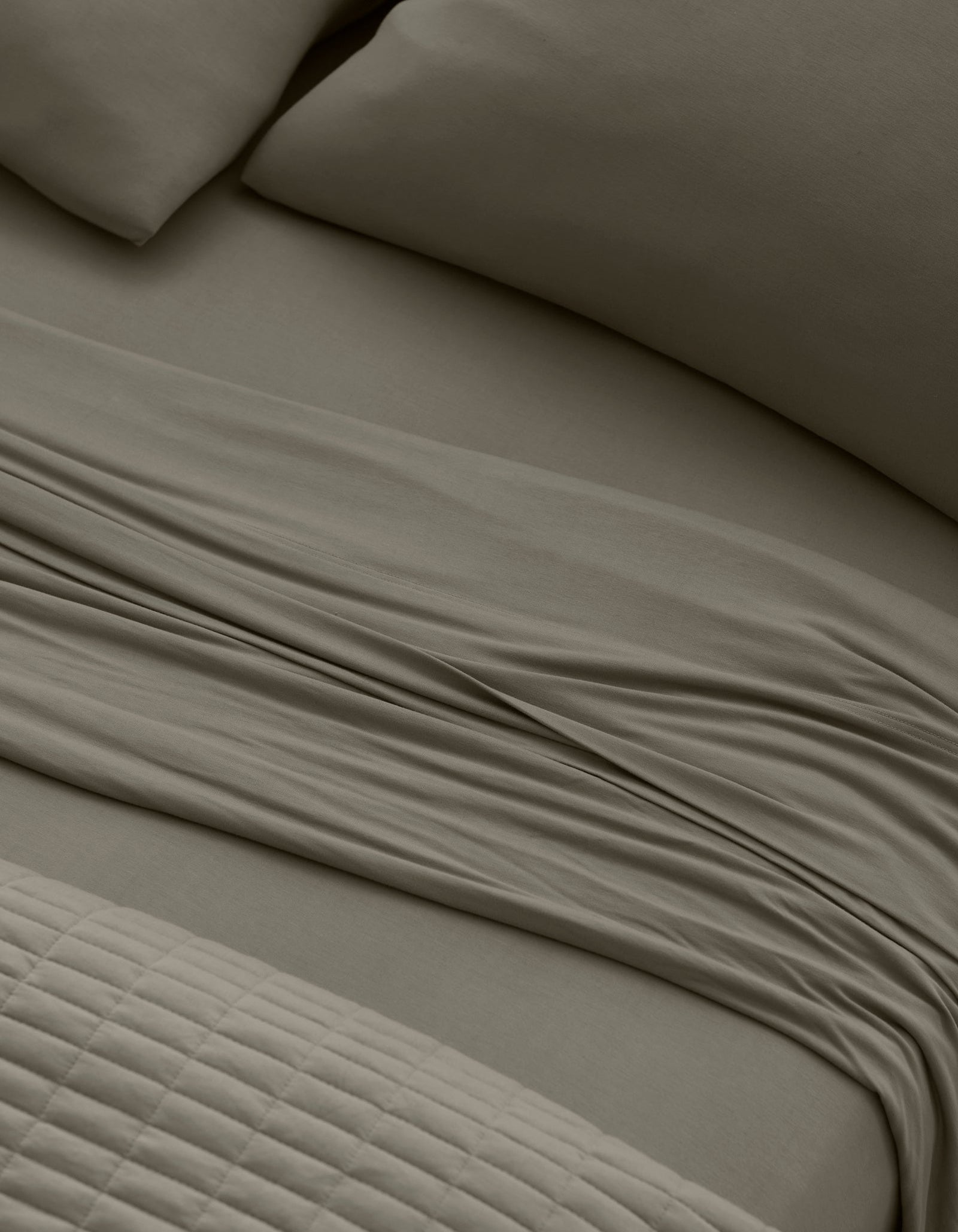 Close-up of a neatly made bed with Cozy Earth Bamboo Jersey Sheet Set in taupe, featuring pillows, a slightly rumpled flat sheet, and a textured quilted blanket at the foot of the bed.
