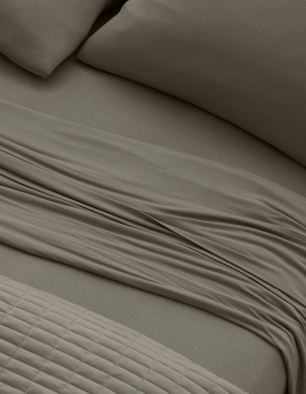 Close-up of a neatly made bed with Cozy Earth Bamboo Jersey Sheet Set in taupe, featuring pillows, a slightly rumpled flat sheet, and a textured quilted blanket at the foot of the bed.|Color:Stone
