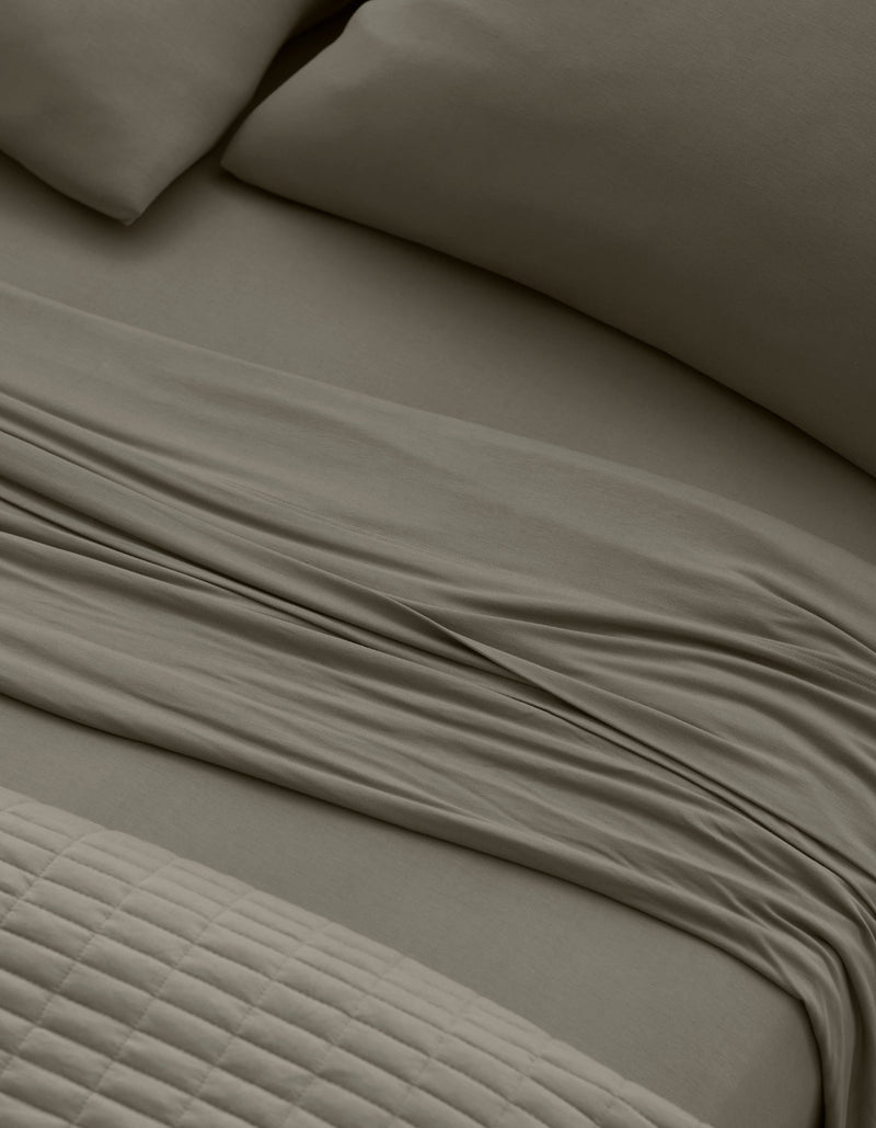 Close-up of a neatly made bed with Cozy Earth Bamboo Jersey Sheet Set in taupe, featuring pillows, a slightly rumpled flat sheet, and a textured quilted blanket at the foot of the bed.