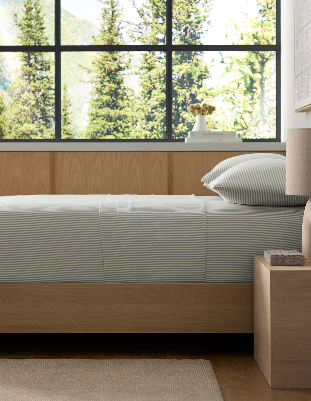 A neatly made bed featuring Cozy Earth’s Bamboo Fitted Sheet sits beside a wooden nightstand and lamp, with large windows framing views of tall evergreen trees and mountains in the background.