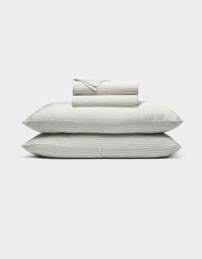 The Bamboo Sheet Set by Cozy Earth features neatly folded white sheets with subtle gray stripes, two stacked pillows, and additional folded sheets, all displayed against a plain white background.|Color:Classic Stripe in Laurel