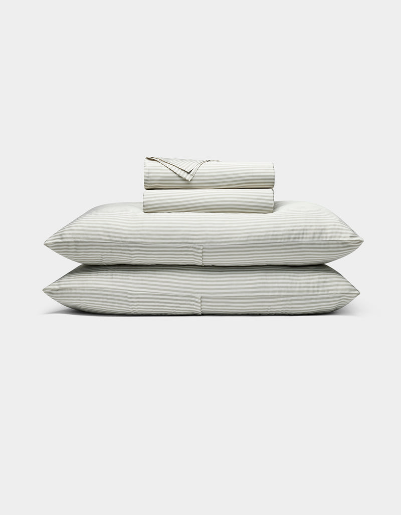 The Bamboo Sheet Set by Cozy Earth features neatly folded white sheets with subtle gray stripes, two stacked pillows, and additional folded sheets, all displayed against a plain white background.