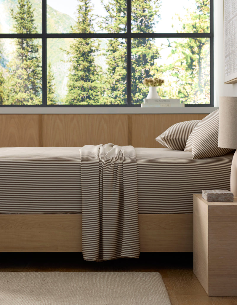 A neatly made bed features Cozy Earth's Bamboo Sheet Set in beige and white stripes, set in a modern bedroom with light wood furniture. Large windows showcase green trees and mountains, filling the room with natural light.