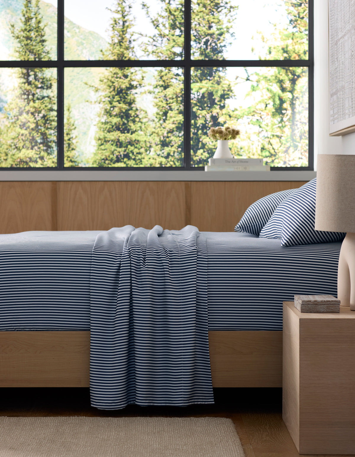 A neatly made bed with Cozy Earth's Bamboo Sheet Set in blue and white stripes sits in a modern bedroom featuring light wood furniture. Large windows frame stunning views of green pine trees and mountains outside.