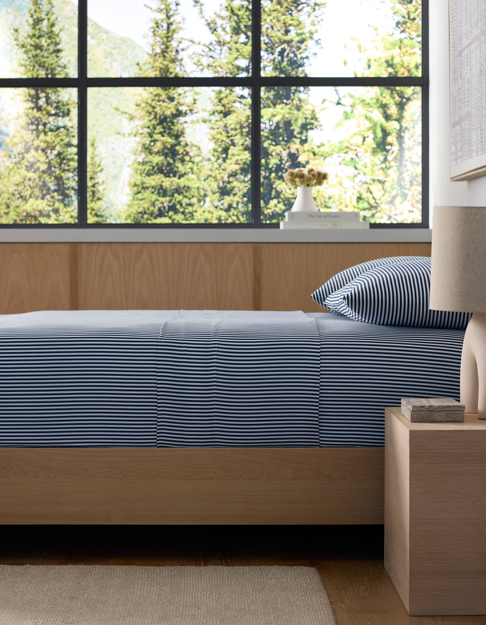 A neatly made bed with Cozy Earth's Bamboo Fitted Sheet is next to a wooden nightstand holding a lamp and book. Large windows offer views of green trees and mountains outside.
