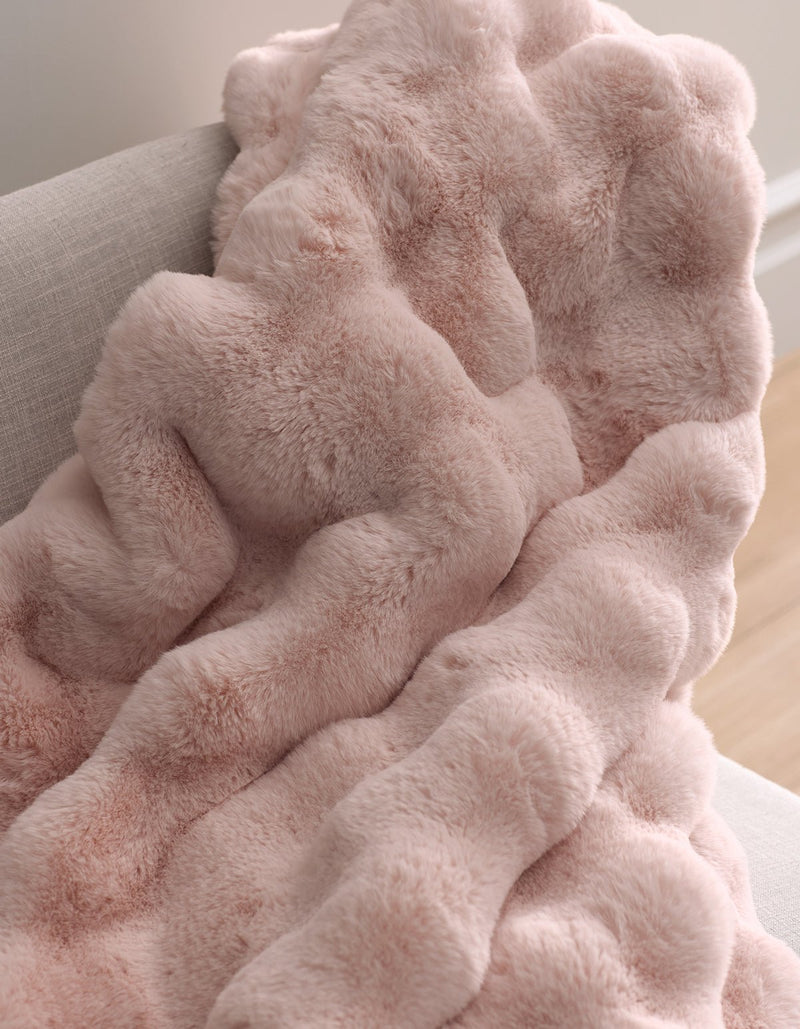 The Bubble Cuddle Blanket by Cozy Earth, featuring soft, plush light pink faux fur with a wavy texture, is draped over the arm of a light gray upholstered chair.