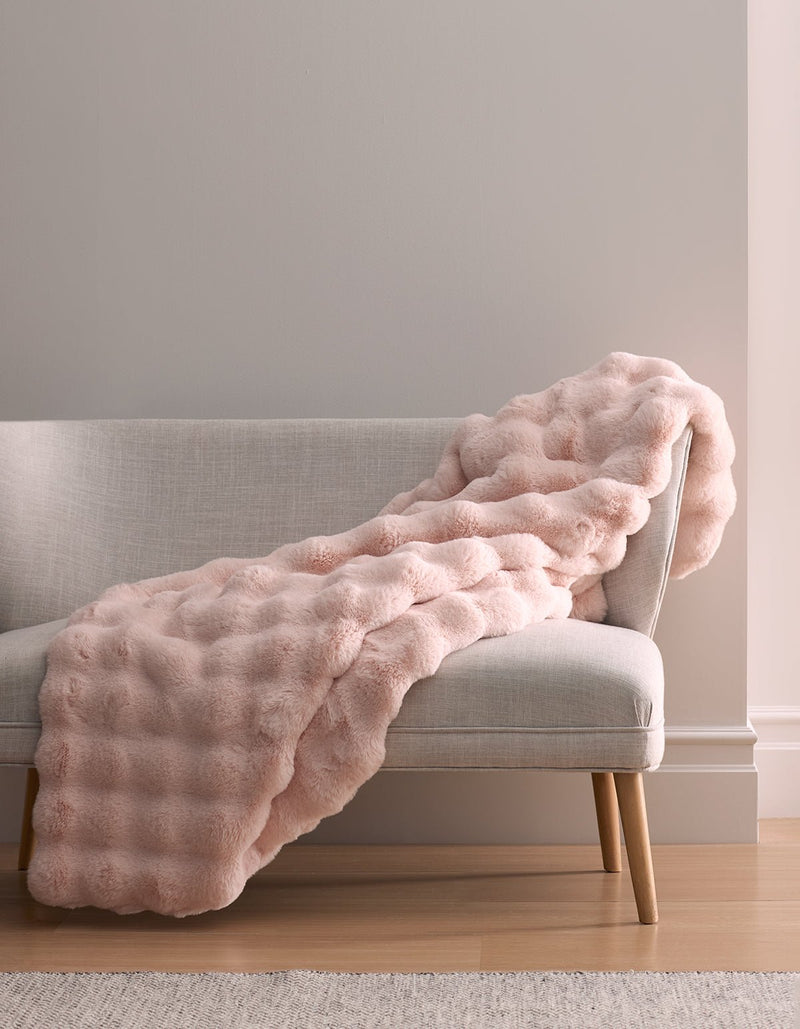 The Bubble Cuddle Blanket by Cozy Earth, in light pink faux fur, is draped over a light gray upholstered bench with wooden legs, against a neutral wall and hardwood floor.