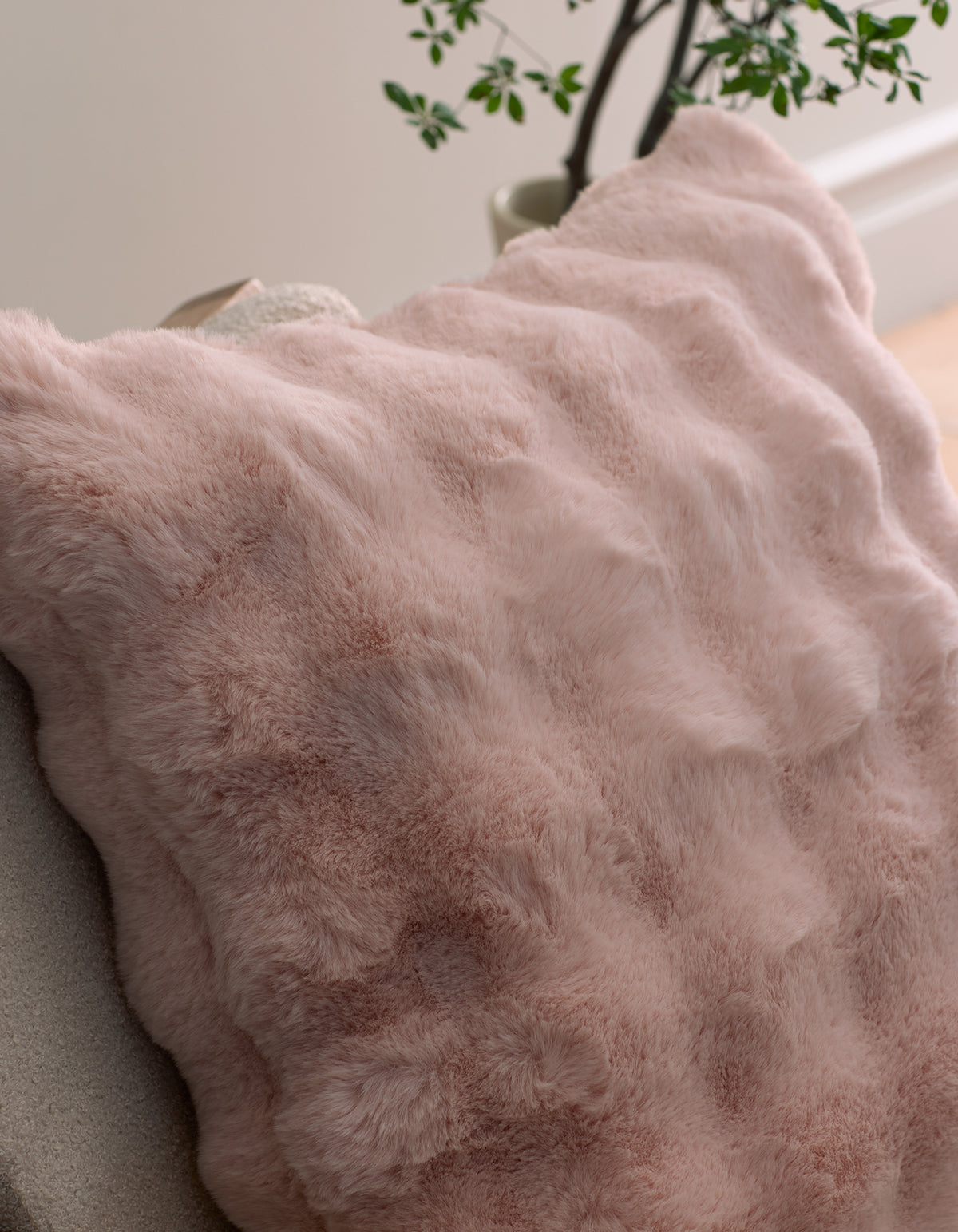 A close-up of the Bubble Cuddle Pillow by Cozy Earth in soft pink faux fur, resting on a chair with a green potted plant visible in the background.