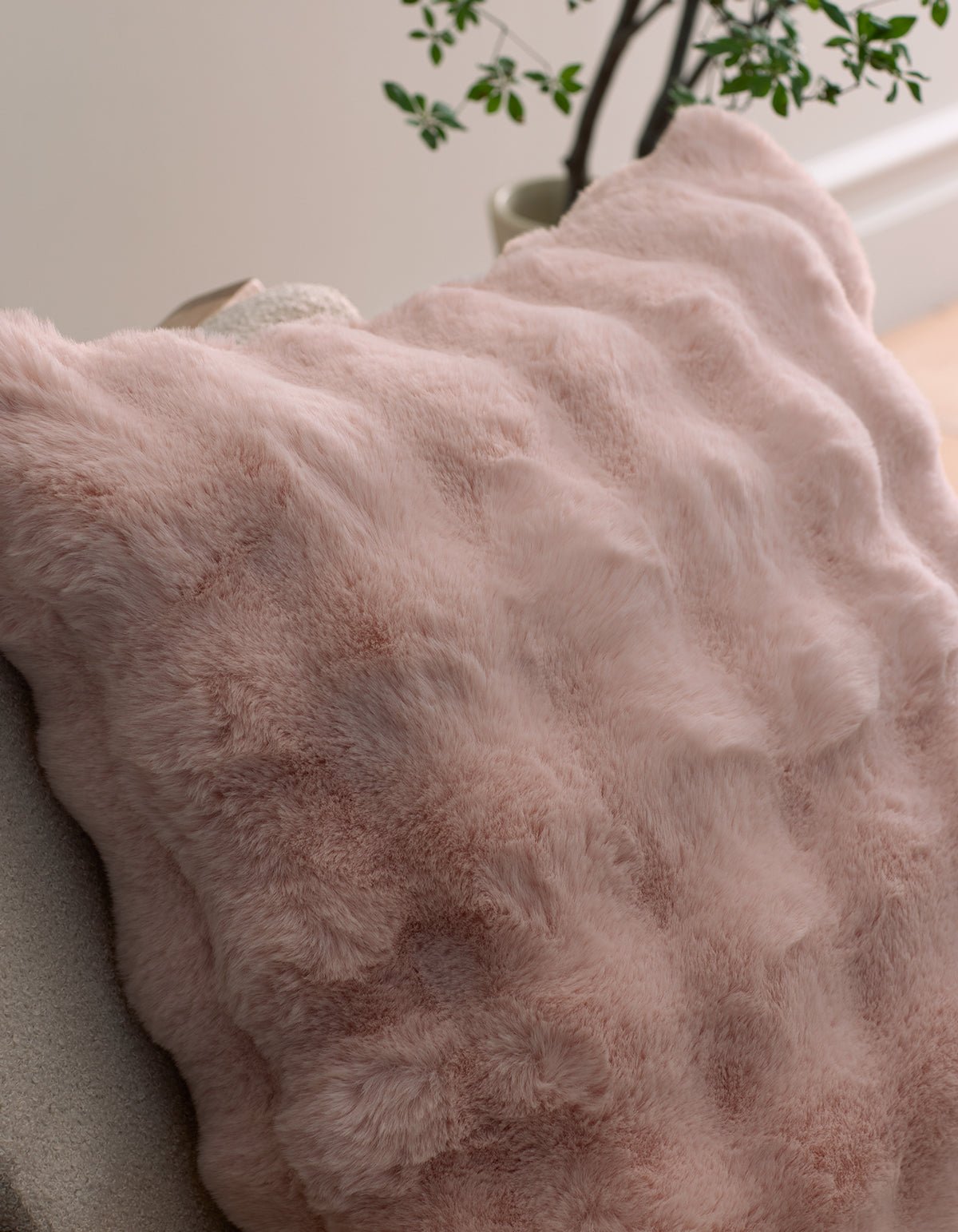 A close-up of the Bubble Cuddle Pillow by Cozy Earth in soft light pink faux fur rests on a chair, with a leafy plant and a pale wall in the background.