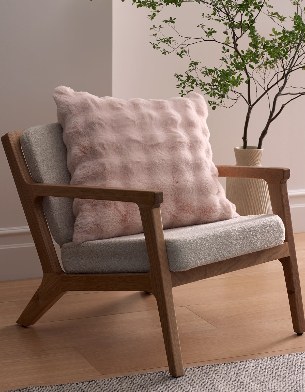A modern wooden armchair with light gray upholstery features the Bubble Cuddle Pillow by Cozy Earth in pink. Nearby, a green leafy plant in a cream vase sits on a light wood floor against a beige wall.